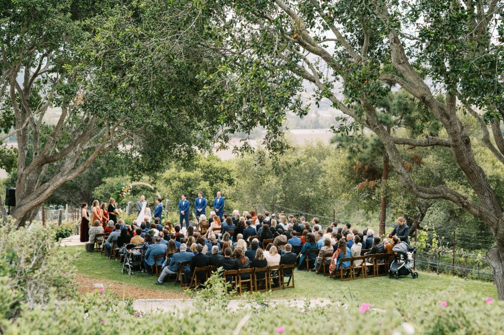 Overview of ceremony in the trees on cloudy day at unique 70s vibe colorful tangerine wedding at Casitas Estate by the best san luis obispo wedding planner Janet Tacy of Tacy and co.