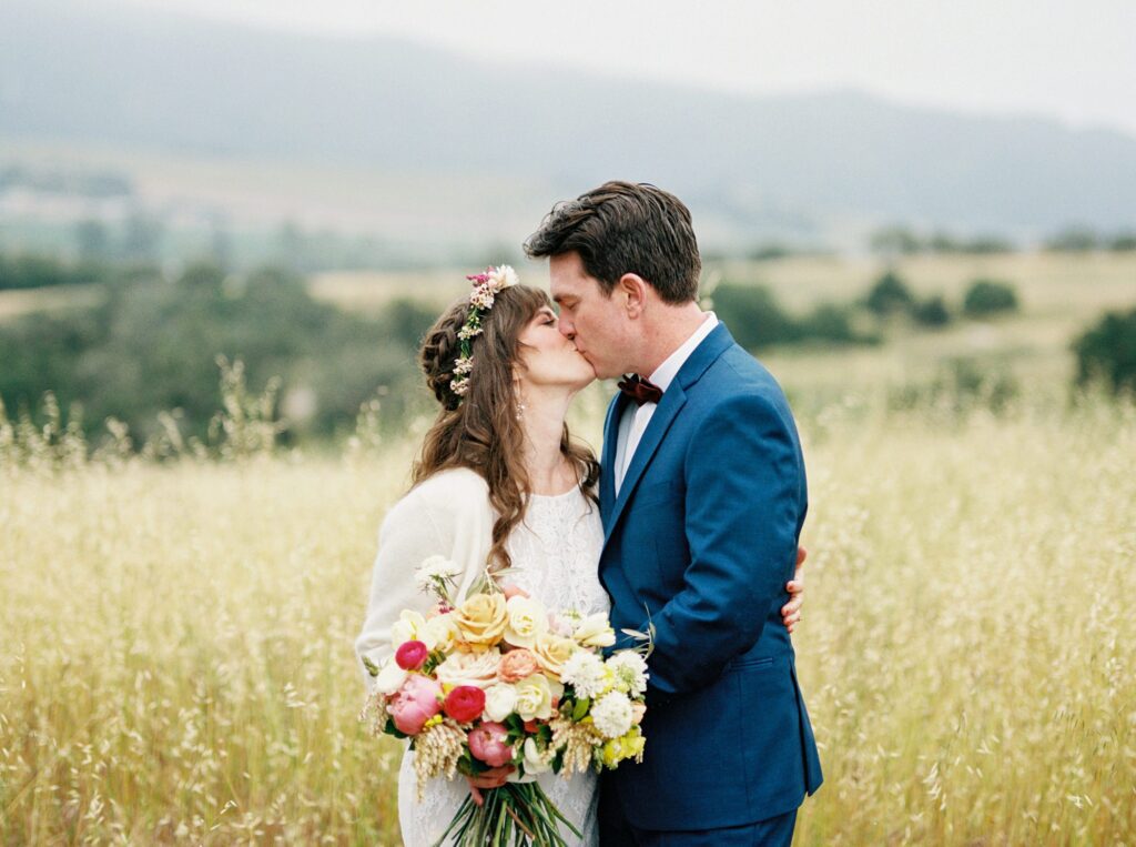 Bride and groom kiss in golden field with hills behind at unique 70s vibe colorful tangerine wedding at Casitas Estate by the best san luis obispo wedding planner Janet Tacy of Tacy and co.